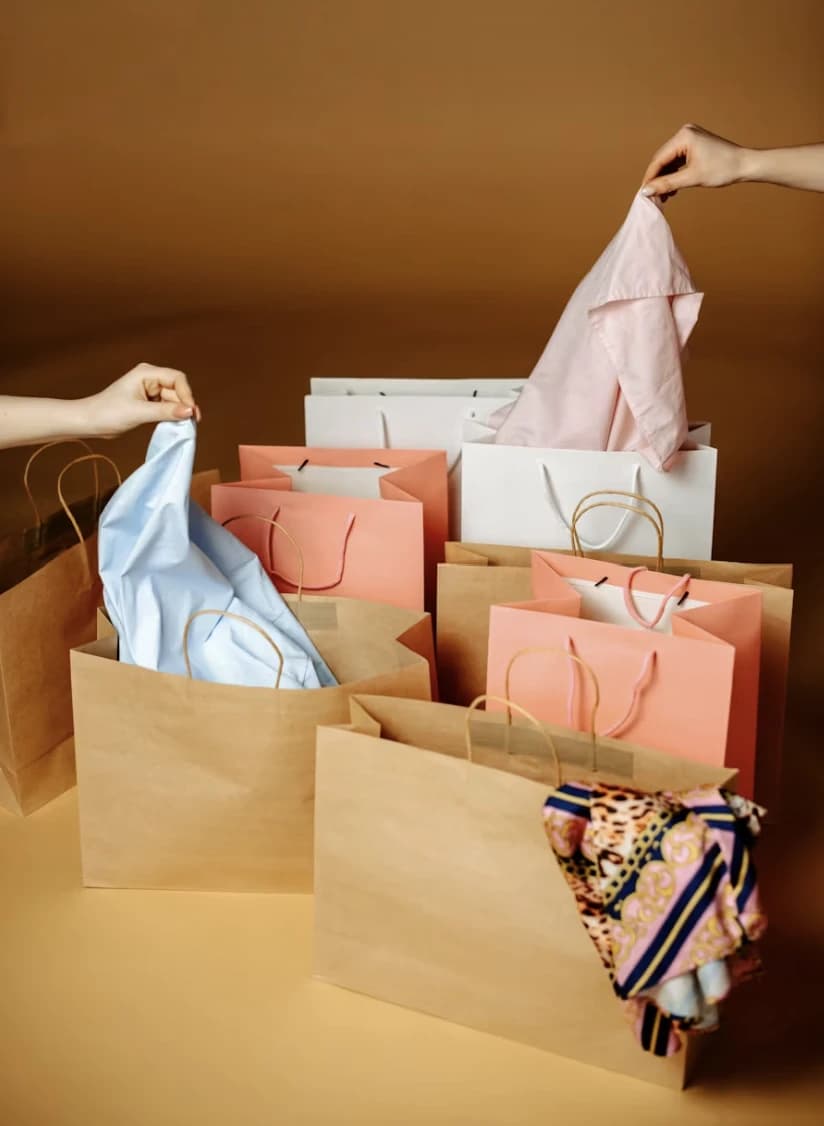 Paper carrier bags of several sizes on a warm ground, folded shirts and a printed scarf being lifted from them.