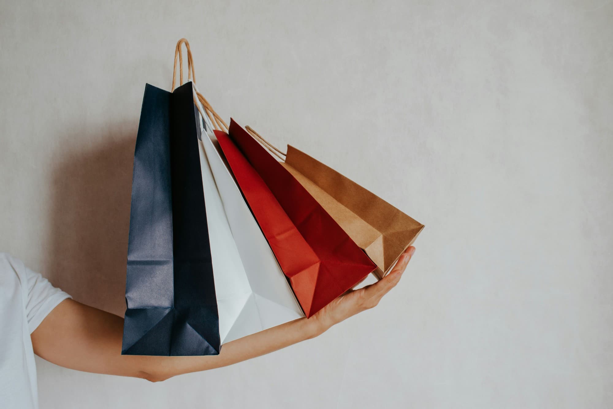 An arm carrying four paper bags by their handles against a plain wall.