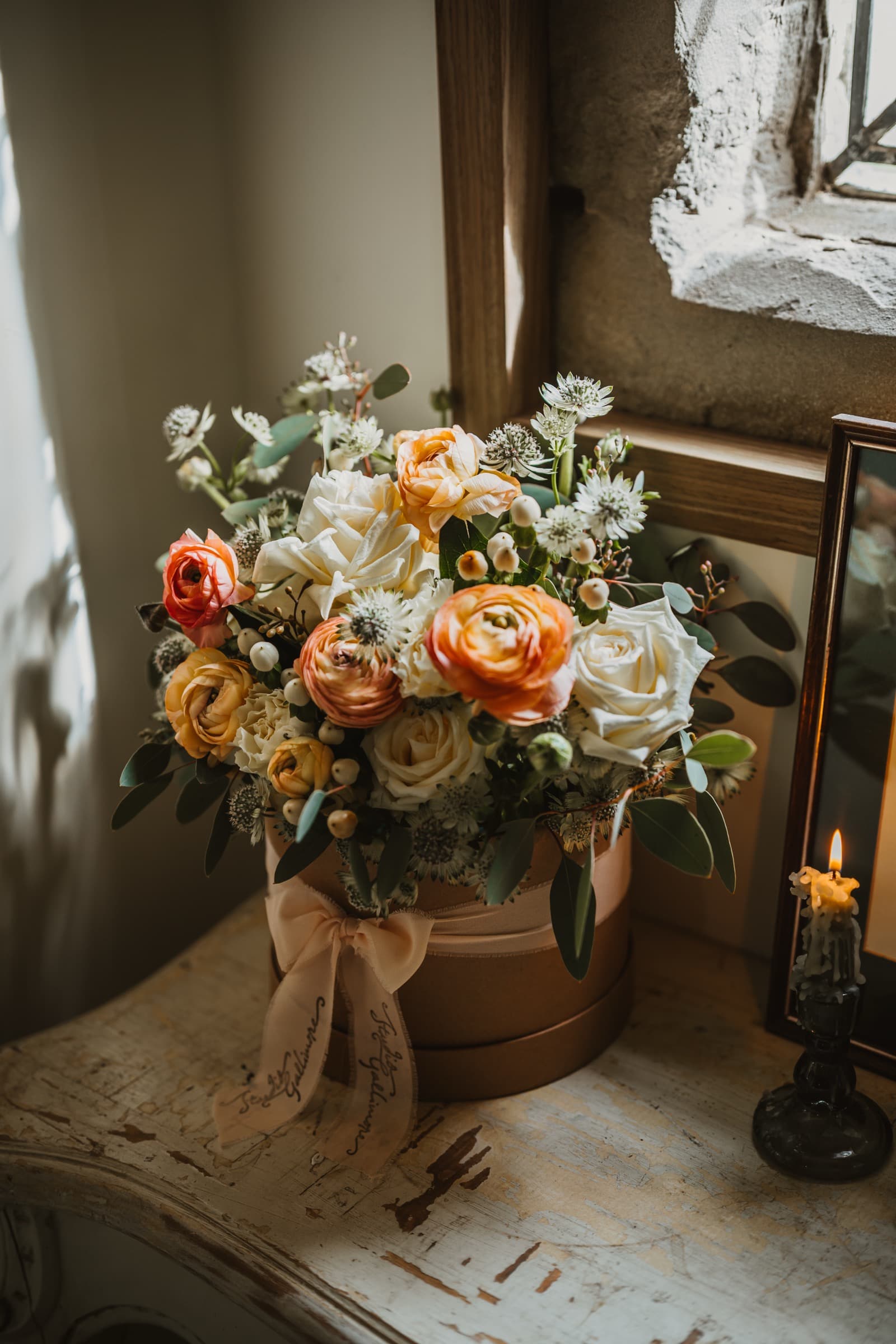 A finished hatbox arrangement of roses, ranunculus and eucalyptus, tied with a Studio Gallimore ribbon, on a painted sill beside a lit candle.