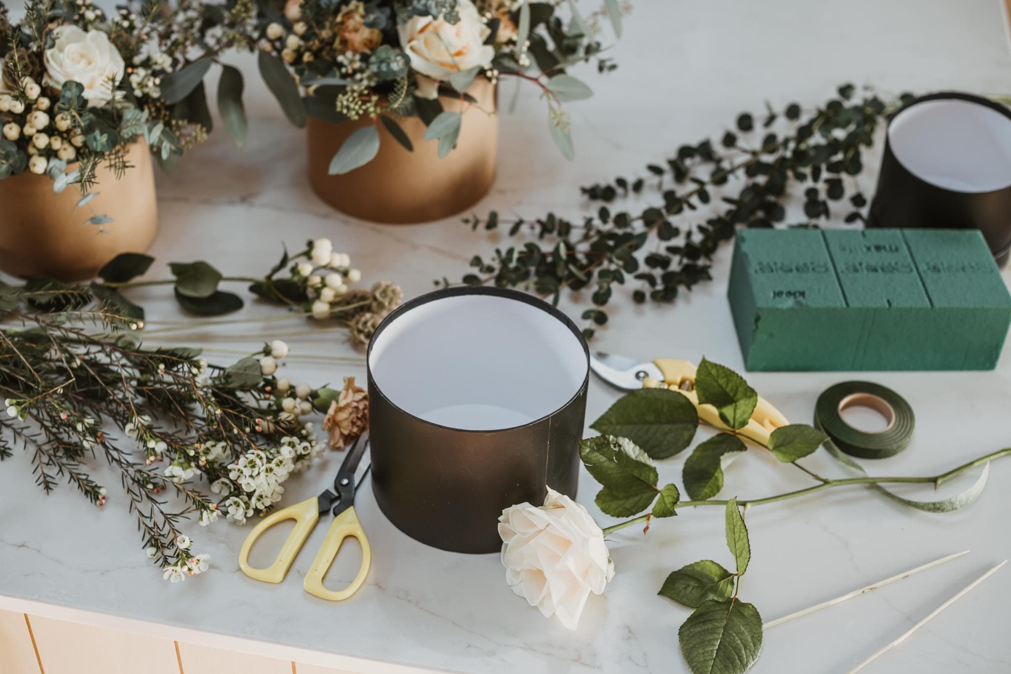 A hatbox, floral foam, secateurs, ribbon and cut stems laid out on a marble surface, with two finished arrangements behind.